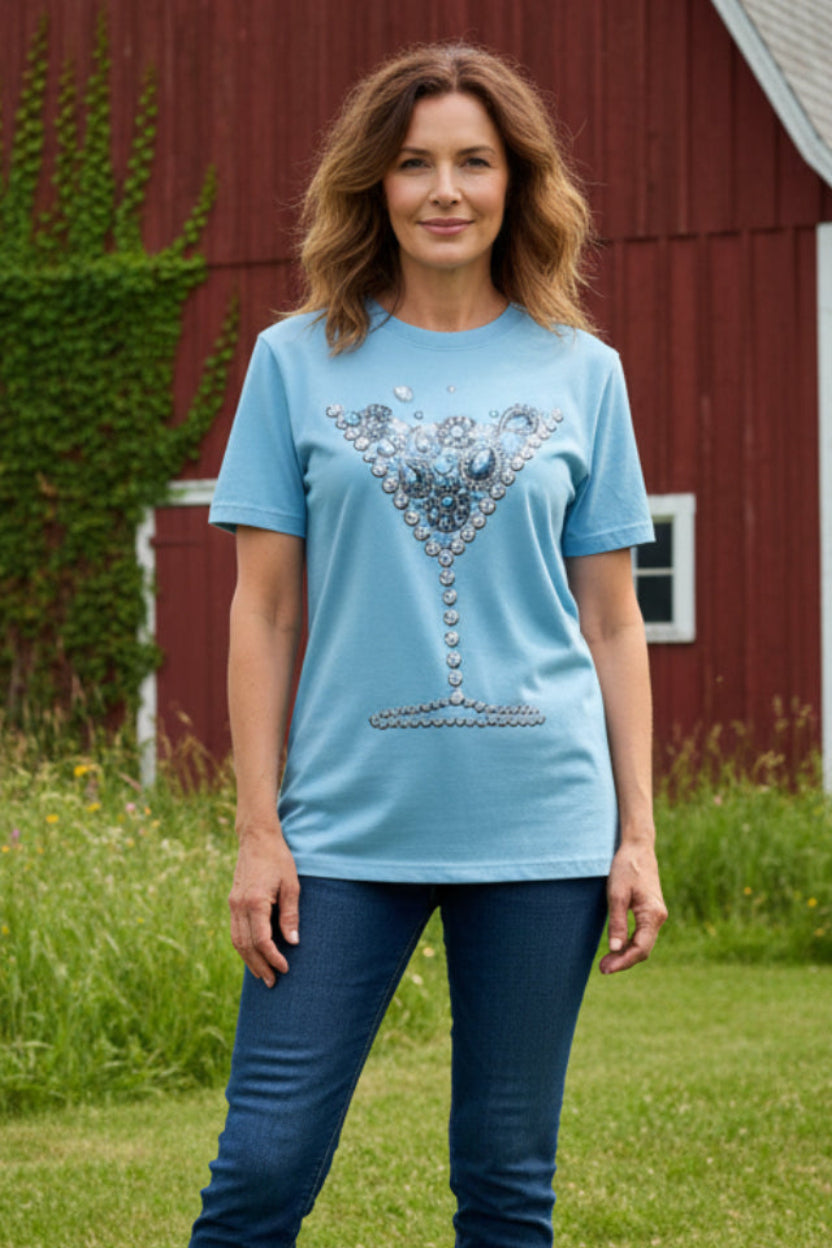 A light blue short-sleeve t-shirt displayed on a mannequin torso in a retail or storage environment. The shirt features a large, detailed graphic of a martini glass filled with silver and blue rhinestone embellishments on the front.