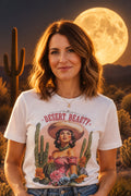 A white short-sleeve graphic t-shirt hanging against a black slatted background. The shirt features a colorful retro-style print of a woman in a sombrero surrounded by cacti and flowers, with the text 'DESERT BEAUTY' and 'Wildwest Western Girl'.