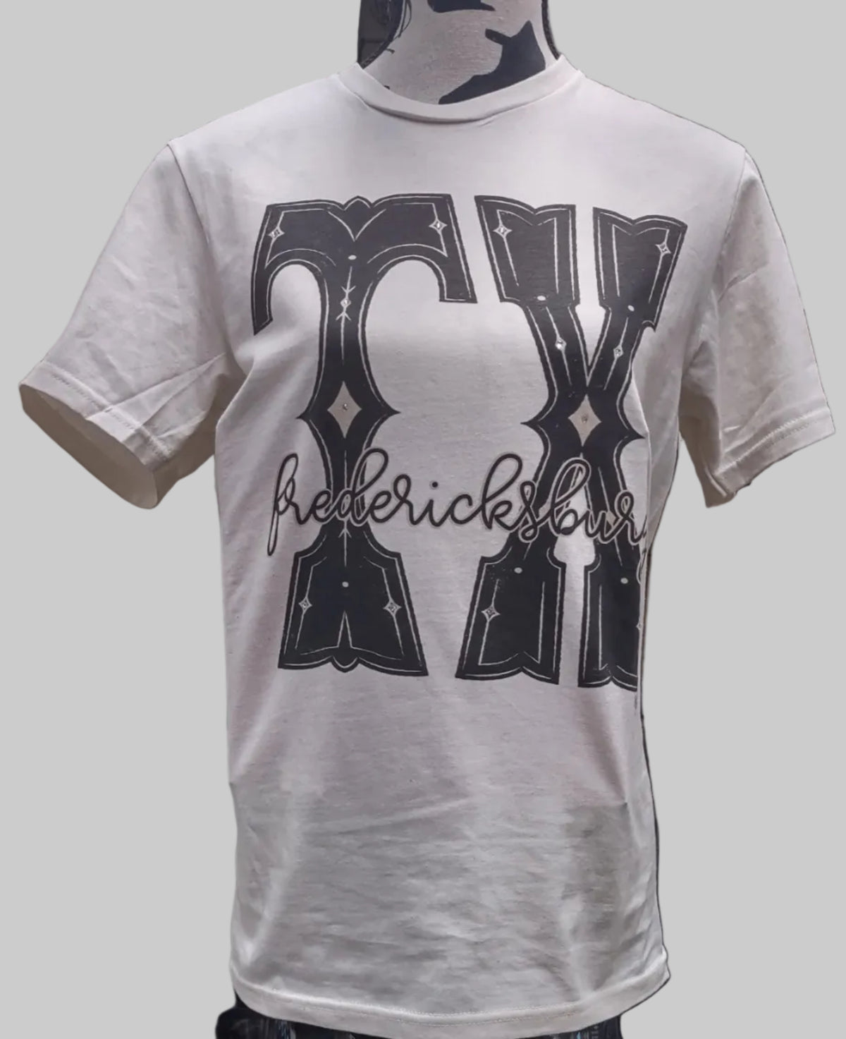 A white short-sleeve T-shirt. The shirt features a large black graphic on the front with the words 'TX' in a stylized font and 'Fredericksburg' written across the chest.