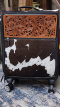 A hard-shell rolling suitcase with a decorative front panel featuring a carved brown leather floral section above a brown-and-white cowhide pattern.