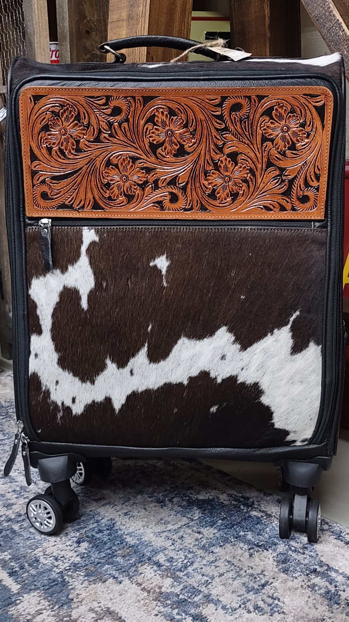 A hard-shell rolling suitcase with a decorative front panel featuring a carved brown leather floral section above a brown-and-white cowhide pattern.