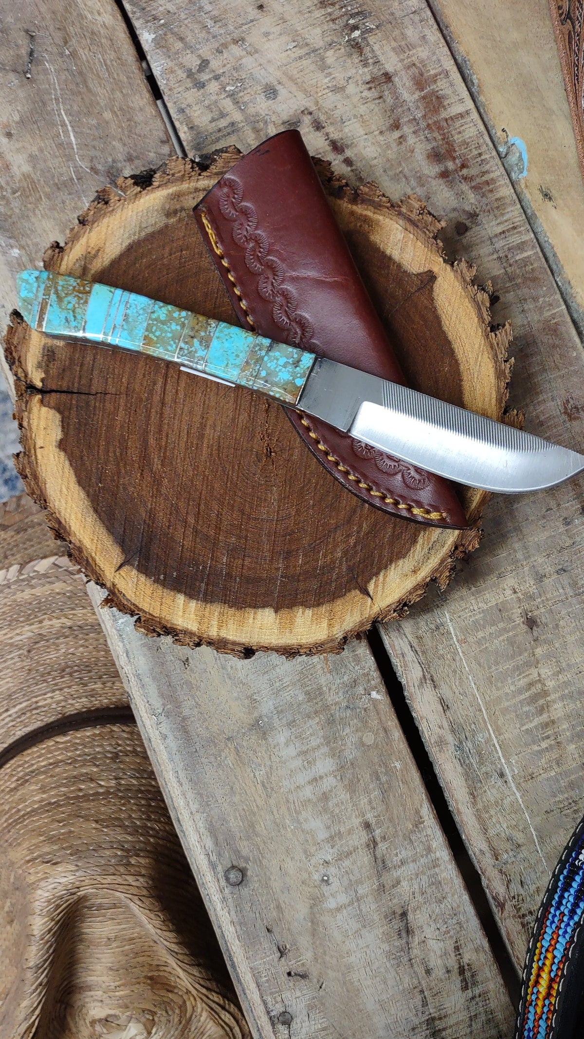 A fixed-blade knife with a turquoise and brown patterned handle and a silver steel blade, resting on a rustic wood slice next to a brown leather sheath, set on a weathered wooden plank.