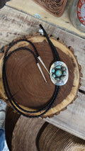 A black braided leather bolo tie with a round silver-toned pendant featuring turquoise stones, displayed on a rustic wood slice over a wooden table.