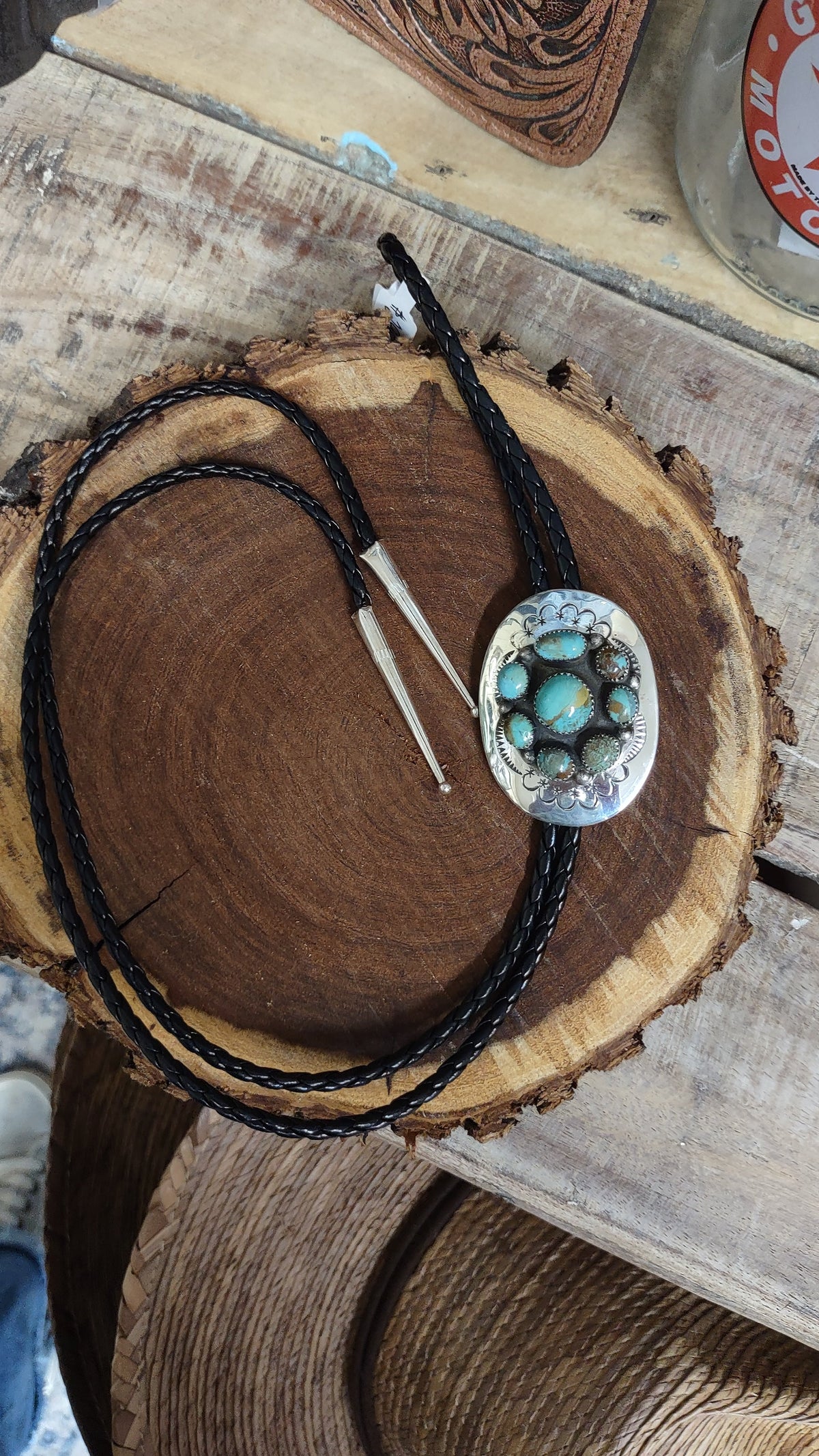 A black braided leather bolo tie with a round silver-toned pendant featuring turquoise stones, displayed on a rustic wood slice over a wooden table.