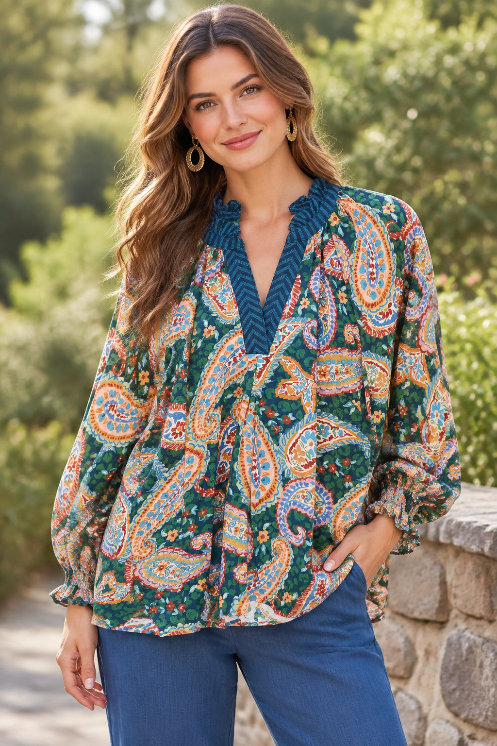  Women's long-sleeve blouse with a vibrant paisley pattern in green, orange, blue, and white, featuring a dark teal chevron-trimmed V-neckline.
